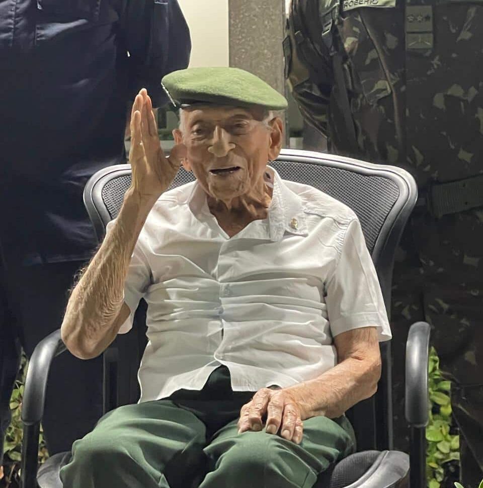 Idoso com boina na cadeira cercado por militares.